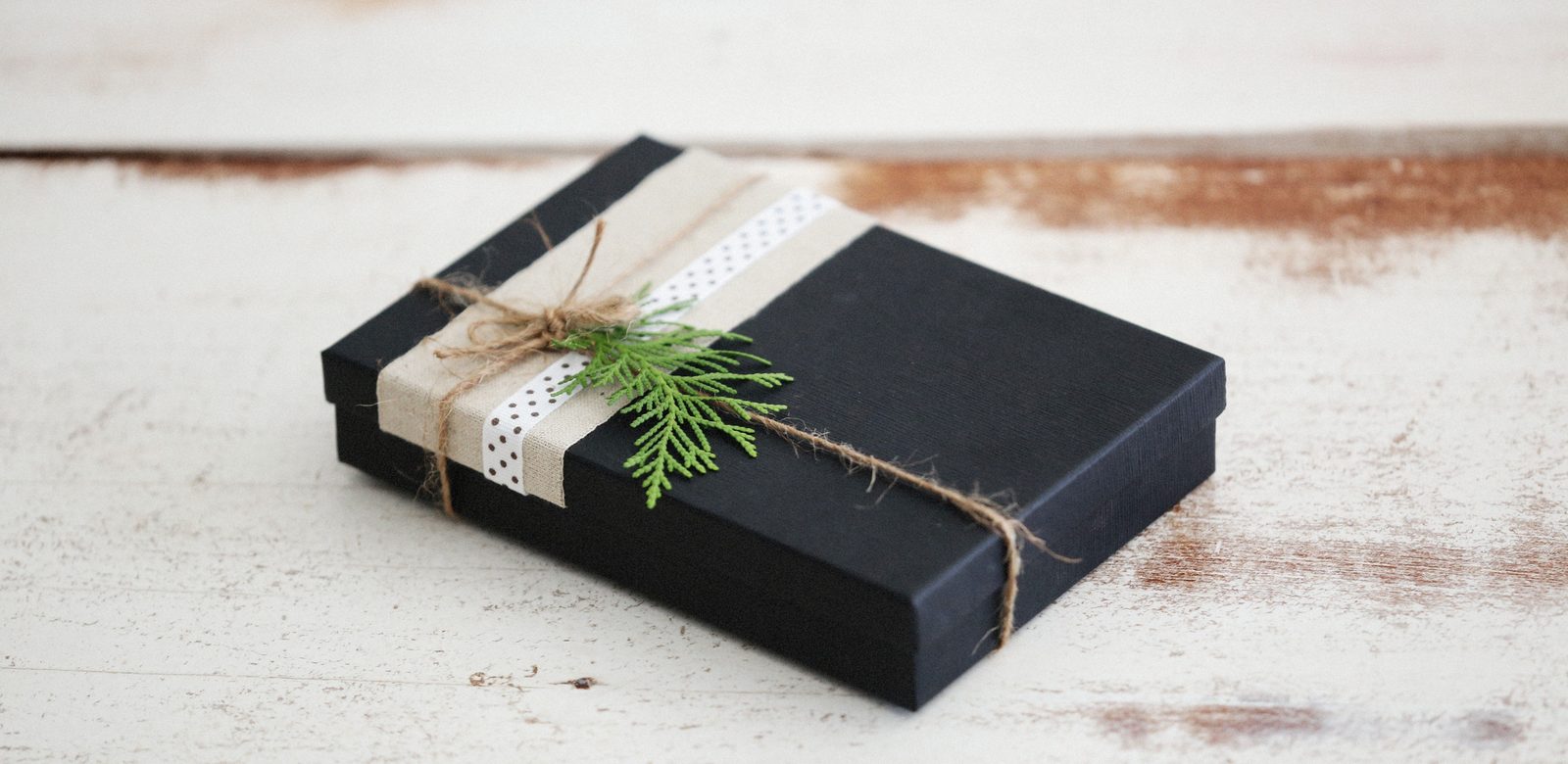 Een zwarte cadeaudoos met een band van linnen, een gestippeld lint, touw en een takje groen op een verweerde witte houten tafel