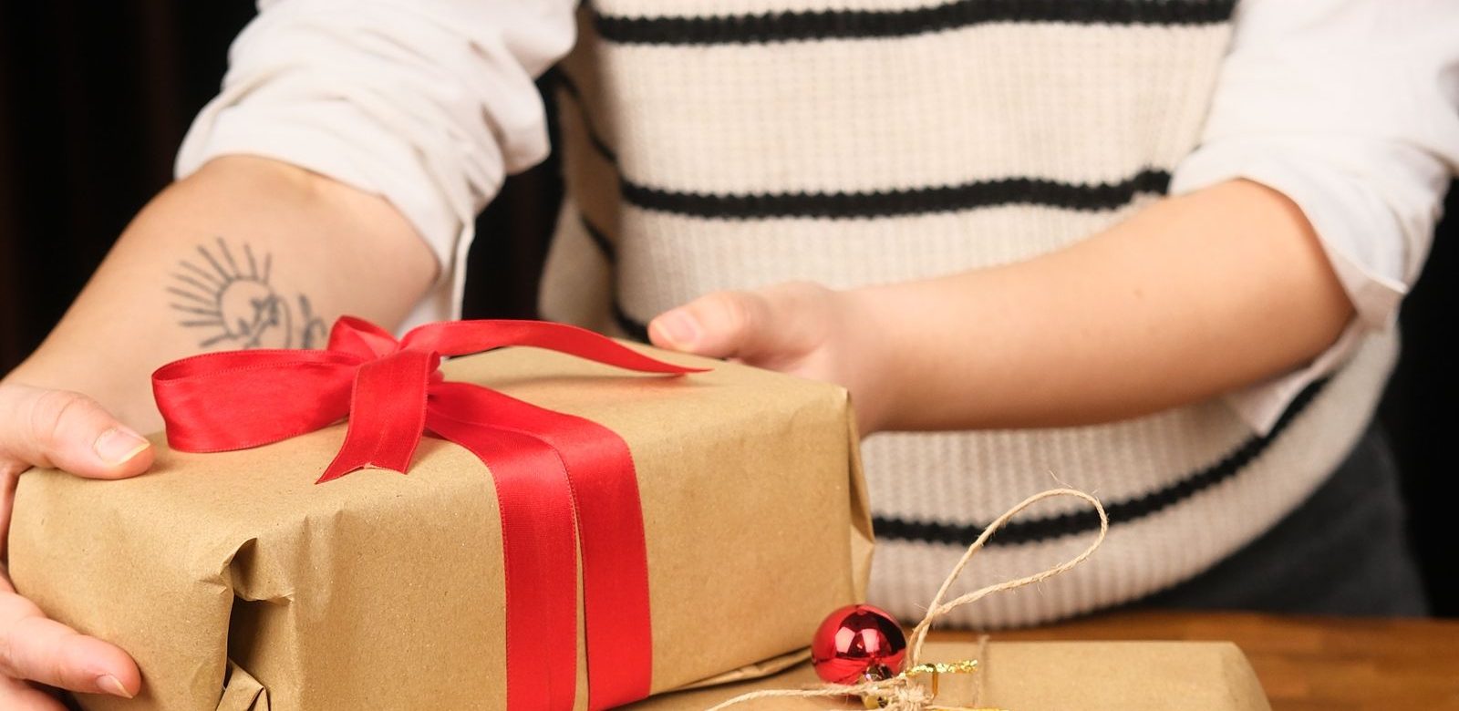 Iemand houdt een cadeau vast dat is ingepakt in bruin papier met een rood lint, met een kerstbal ernaast
