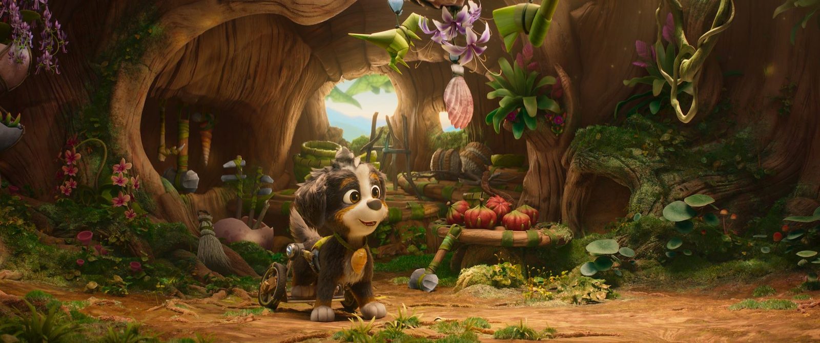 Pup Rex staat in een grot vol planten en bloemen naast een kar met fruit, officieel persbeeld uit Paw Patrol: De Dinofilm