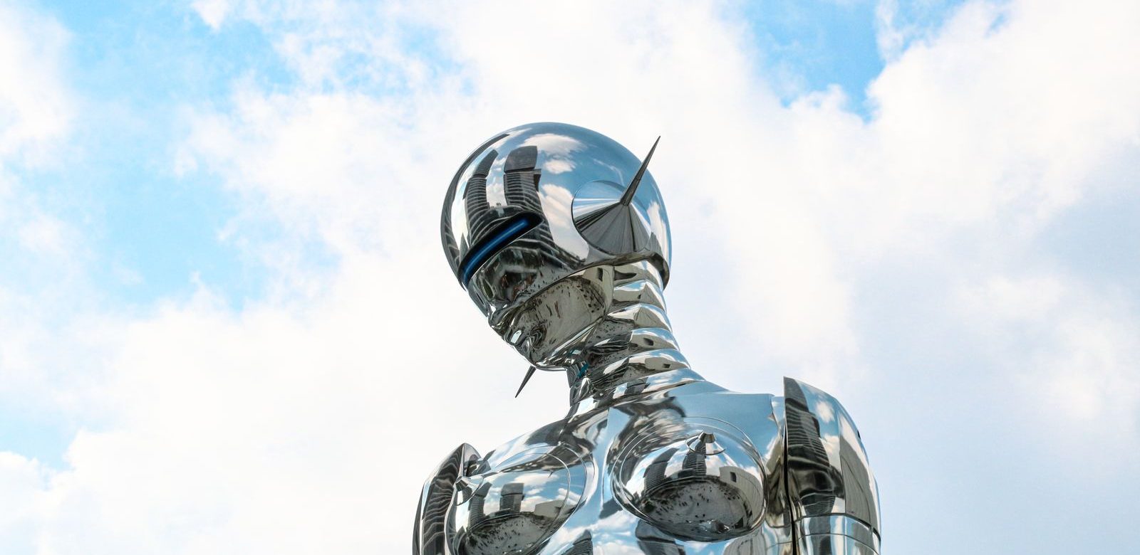 Een glanzend chromen beeld van een figuur met een helm, tegen een blauwe lucht met wolken