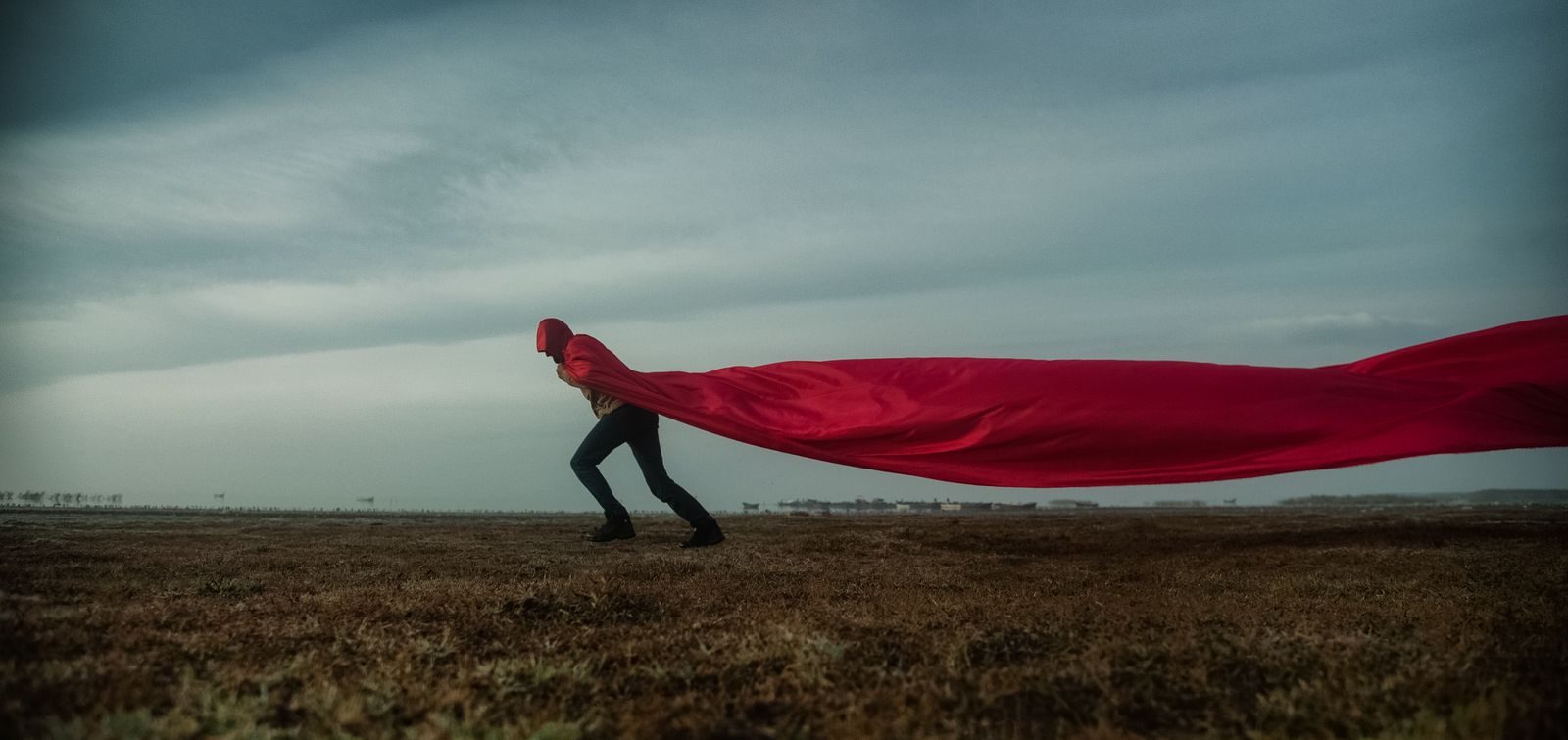 Iemand in een lange rode cape die in de wind uitwaait, staand in een leeg veld onder een grijze lucht