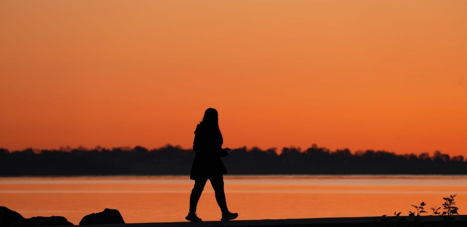 Het silhouet van een wandelende vrouw langs het water bij een oranje zonsondergang