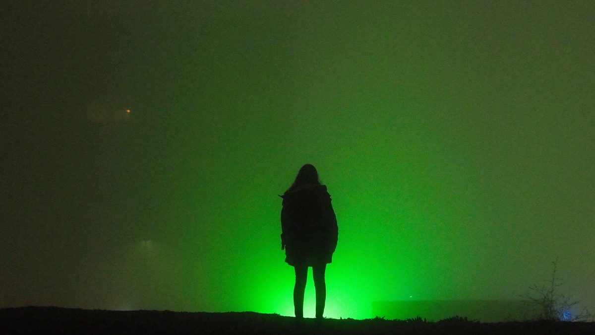 Een silhouet van een persoon in het donker, voor een fel groen licht in de mist