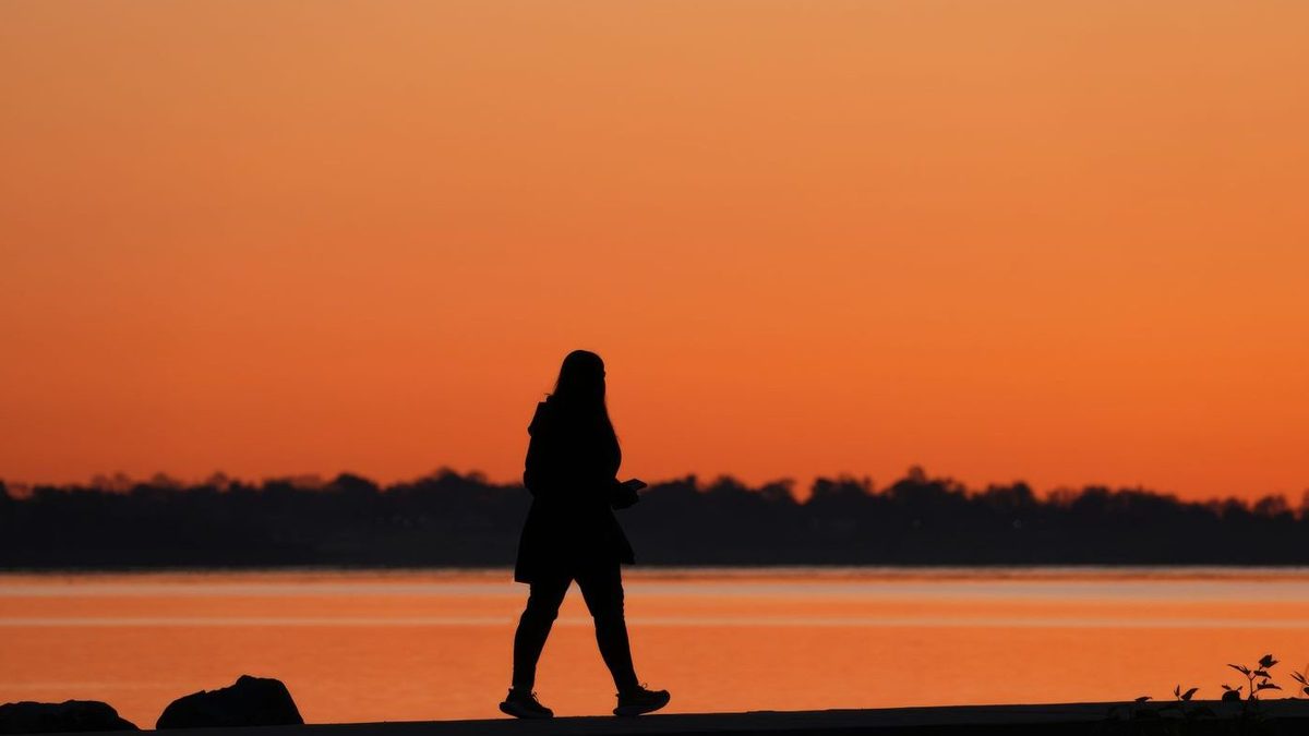 Het silhouet van een wandelende vrouw langs het water bij een oranje zonsondergang