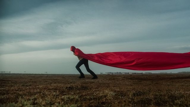 Iemand in een lange rode cape die in de wind uitwaait, staand in een leeg veld onder een grijze lucht