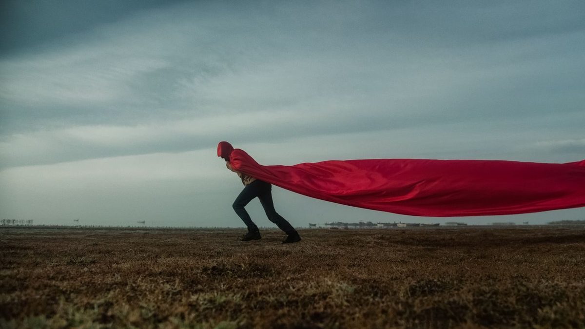 Iemand in een lange rode cape die in de wind uitwaait, staand in een leeg veld onder een grijze lucht